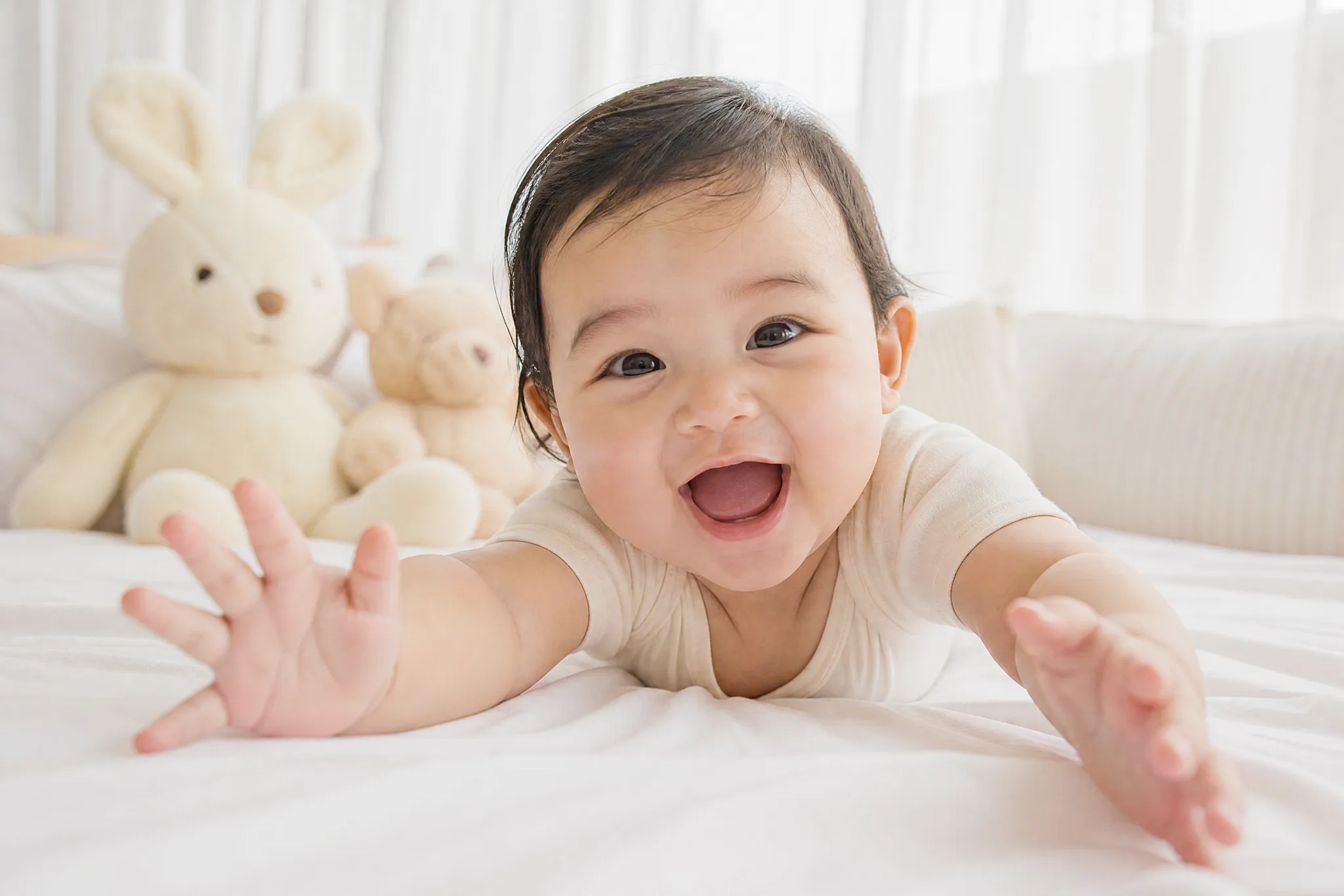 Laughing baby on its tummy reaching both hands toward the camera on a white bed, soft toys and window light behind