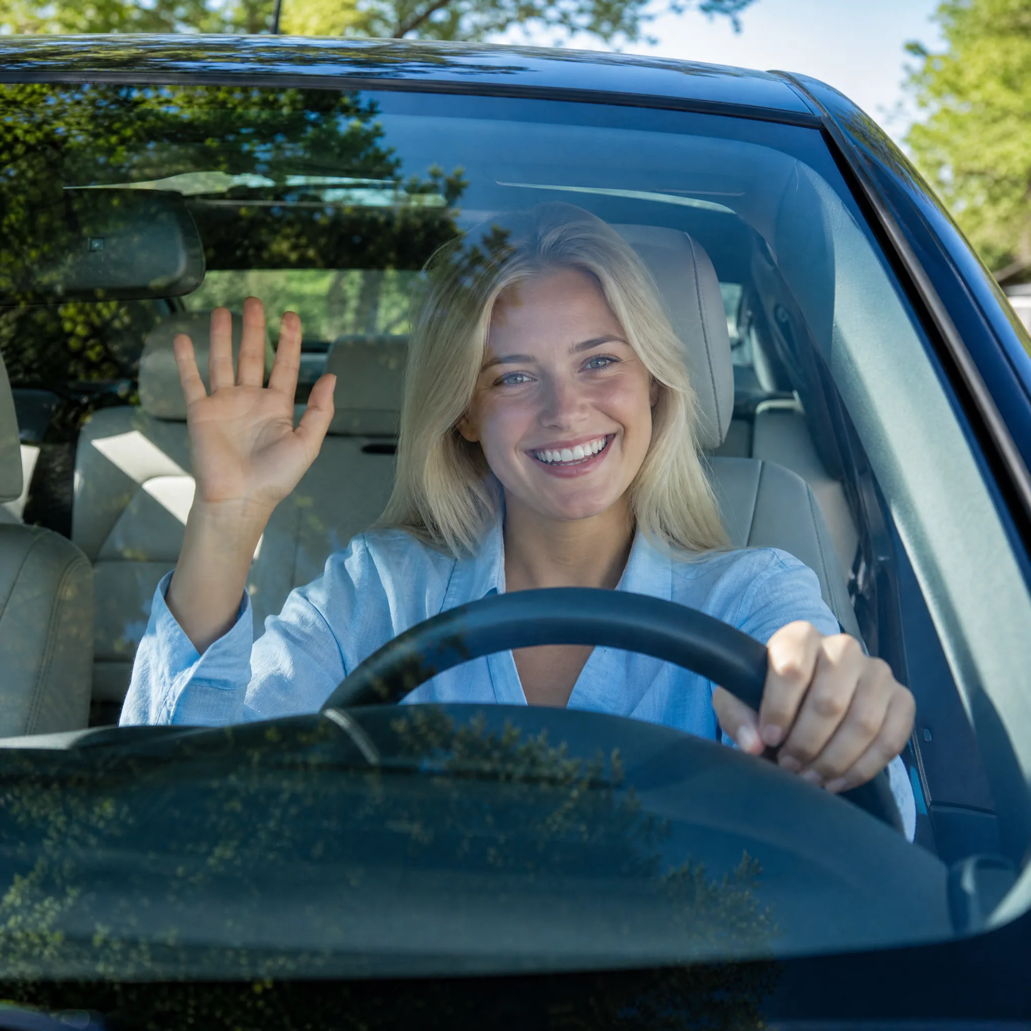 Platinum-blonde woman in a pale blue linen shirt laughing and waving through a windshield, green trees reflected in the glass