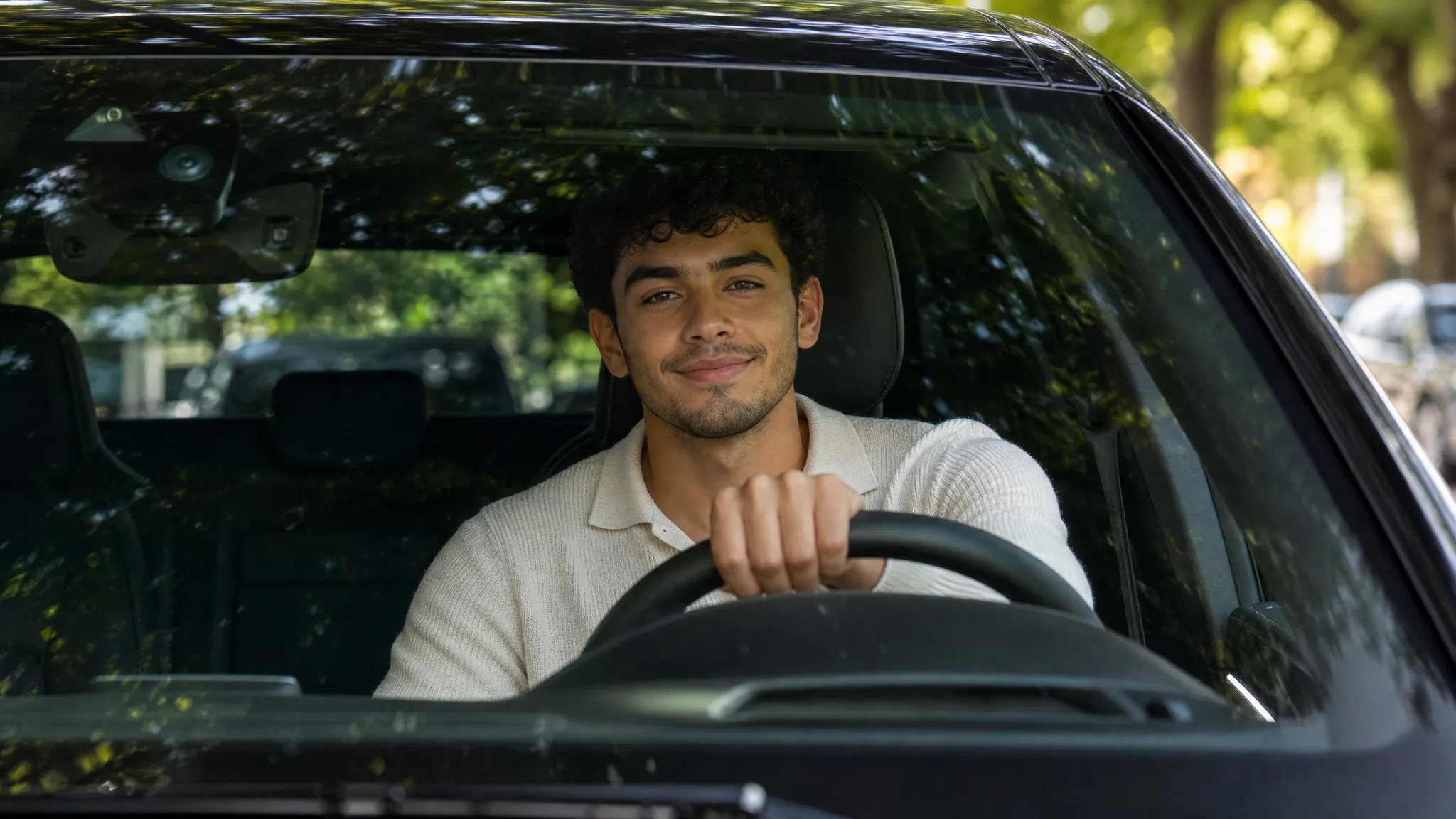 AI Car Video Generator: A Headshot Behind the Wheel Curly-haired young man in a cream knit polo smiling through a car windshield, one hand on the wheel under leafy light