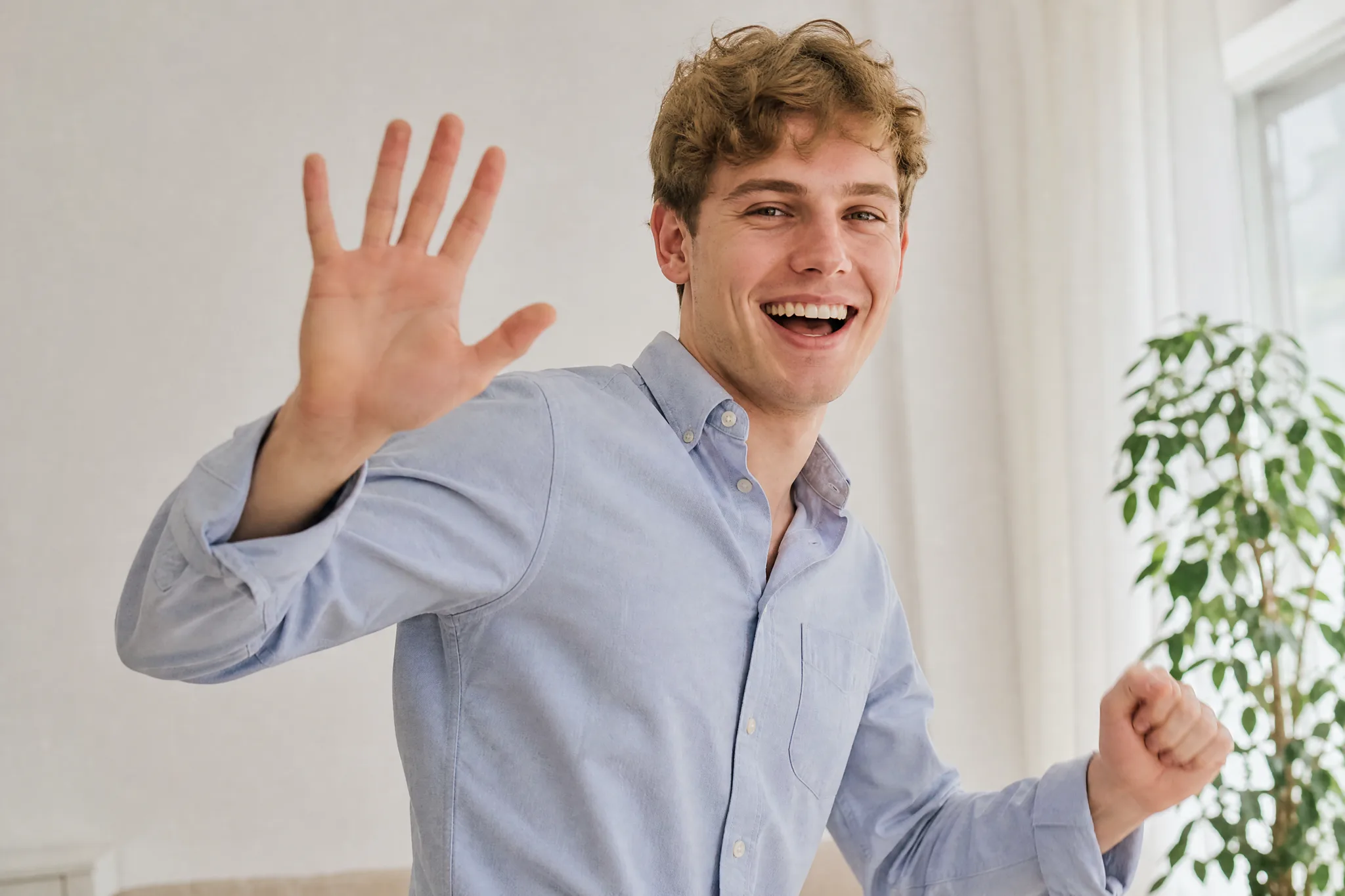 Laughing young man with wavy light brown hair waving one open hand at the camera in a bright room beside a window