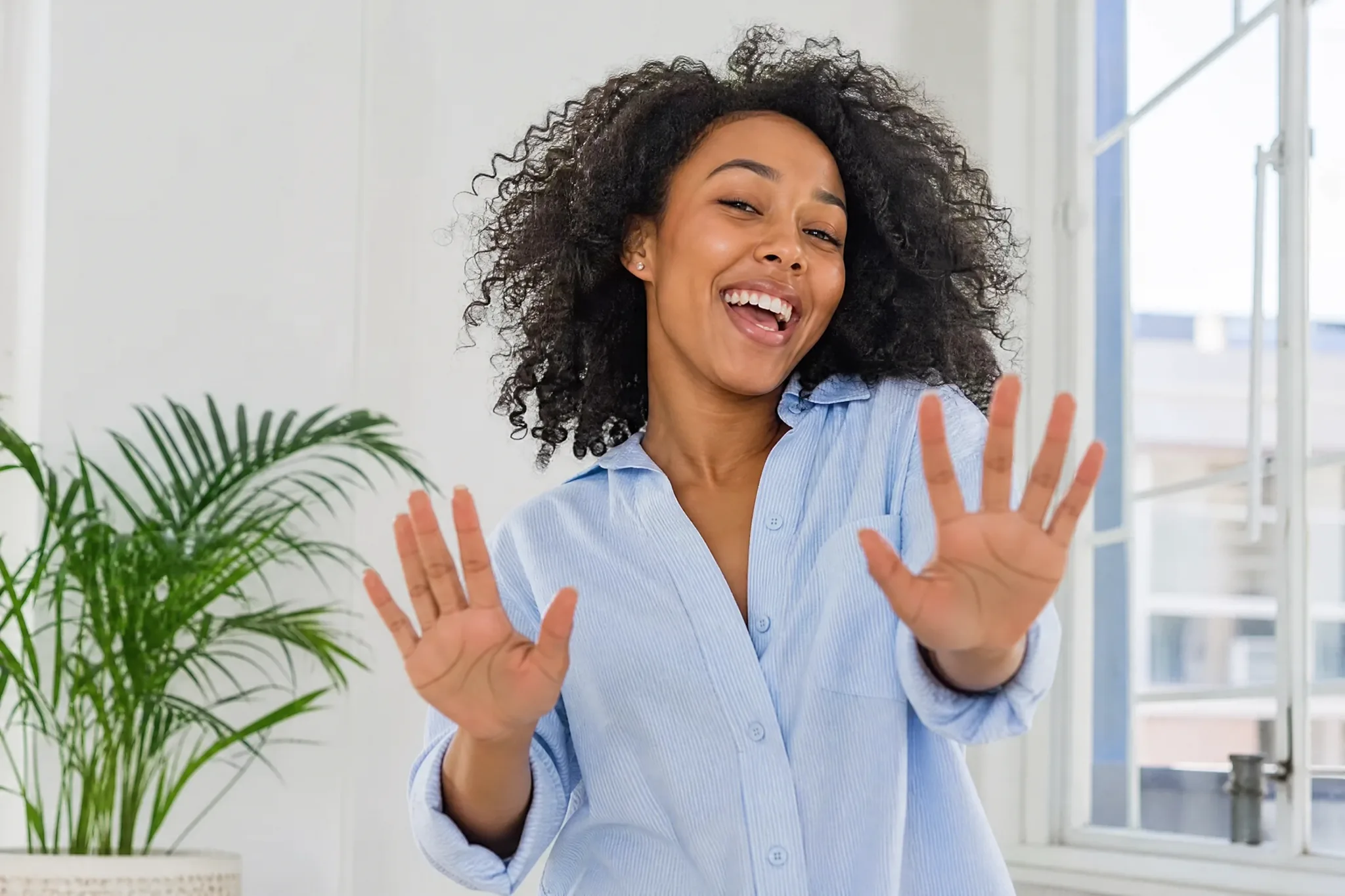 Curly-haired young woman in a light blue shirt holding both palms up to the camera with a wide grin in a white room