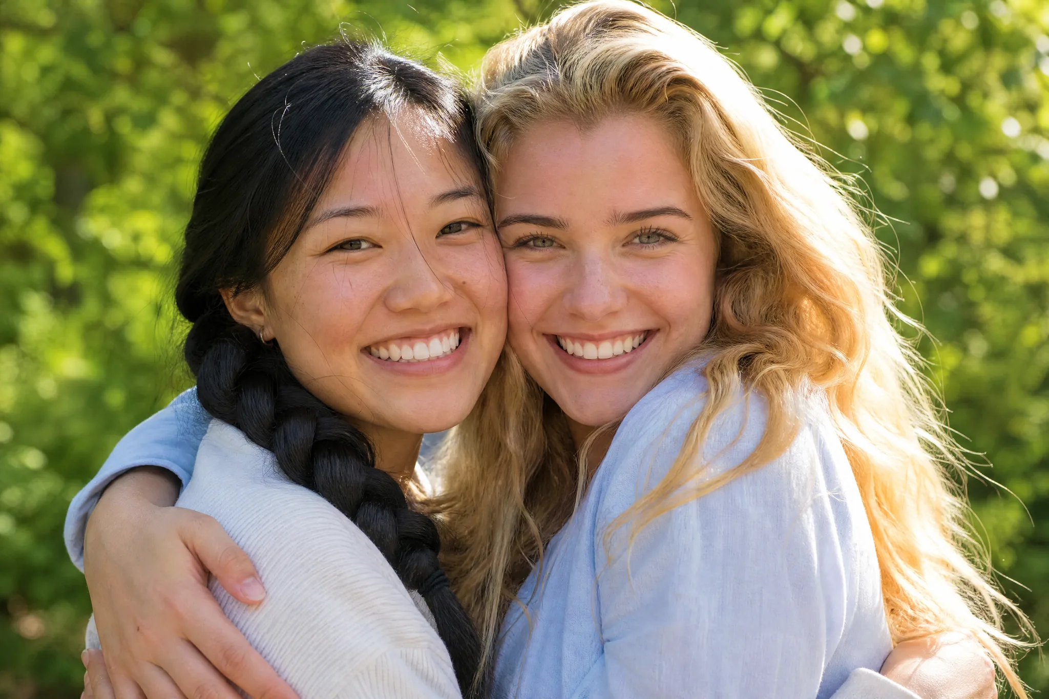 AI Hugging Video: Cheek to Cheek Two young women smiling cheek to cheek in the middle of a hug, one with a black braid, green trees in the afternoon sun
