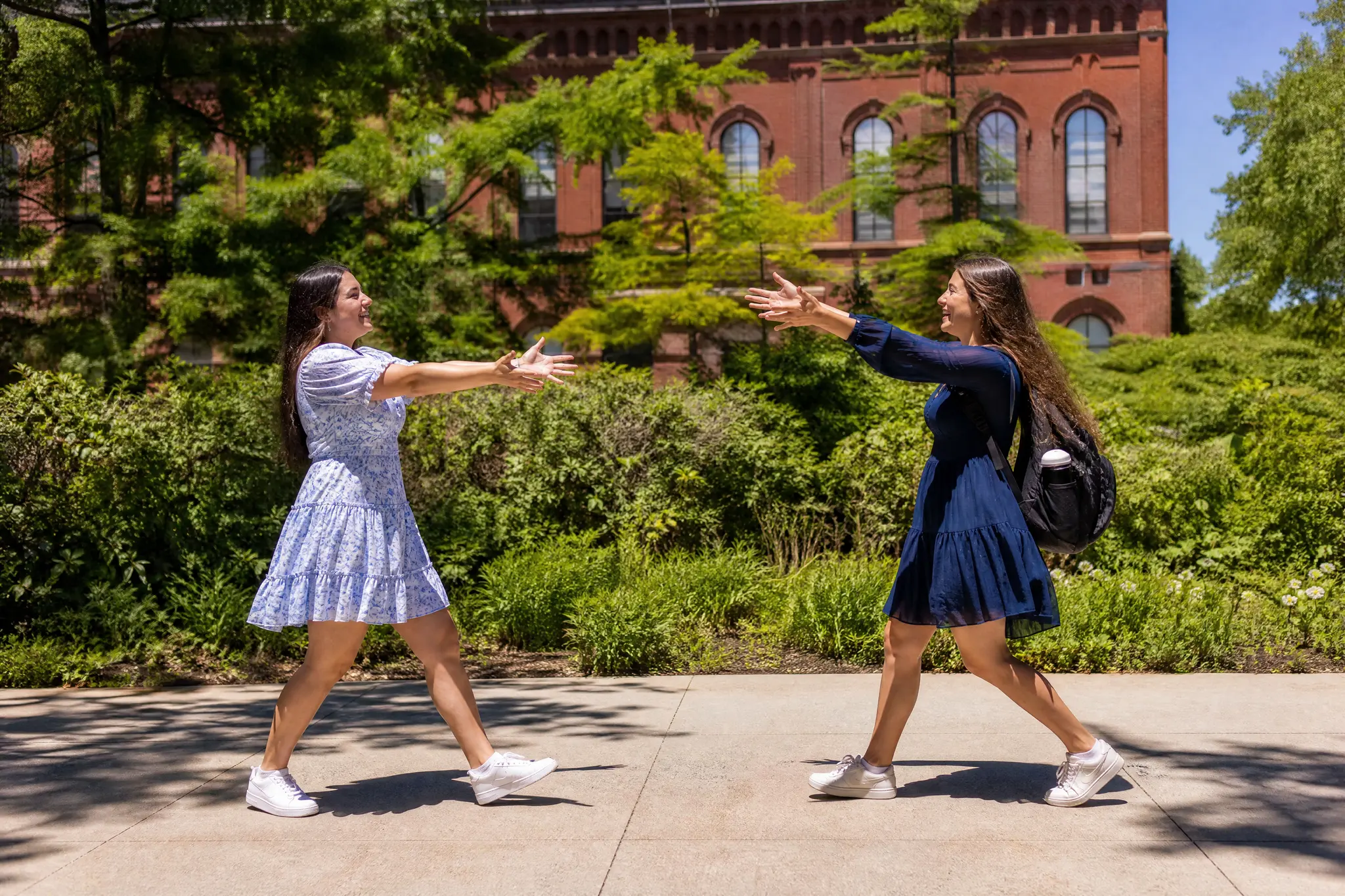 Hugging Video AI: Left Friend, Right Friend Side view of two young women in blue floral and navy dresses walking toward each other with open arms by a brick building