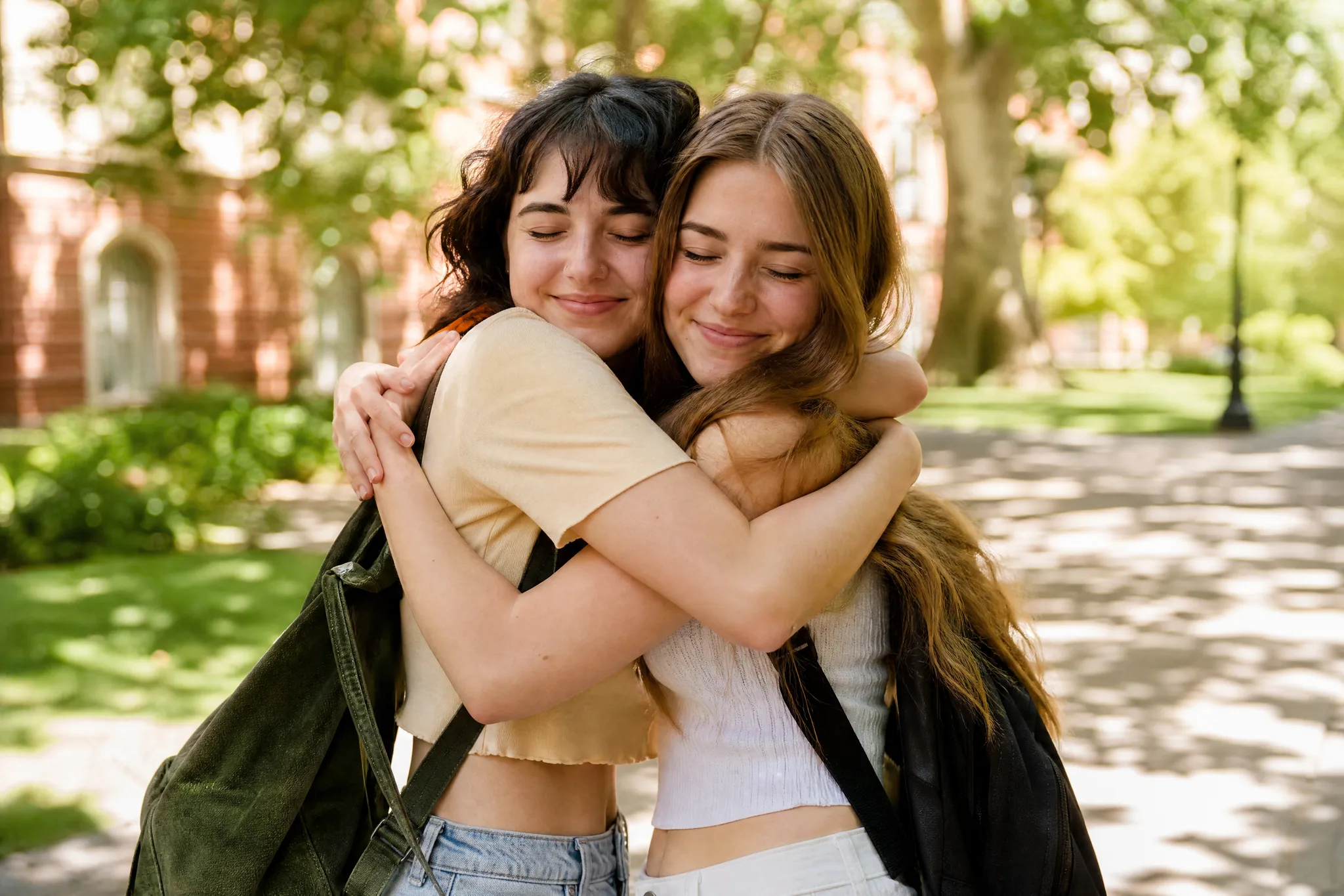 AI Hugging Video: One Big Hug from Two Photos Two young women with eyes closed holding each other in a big hug on a sunlit walkway, trees and red brick blurred behind