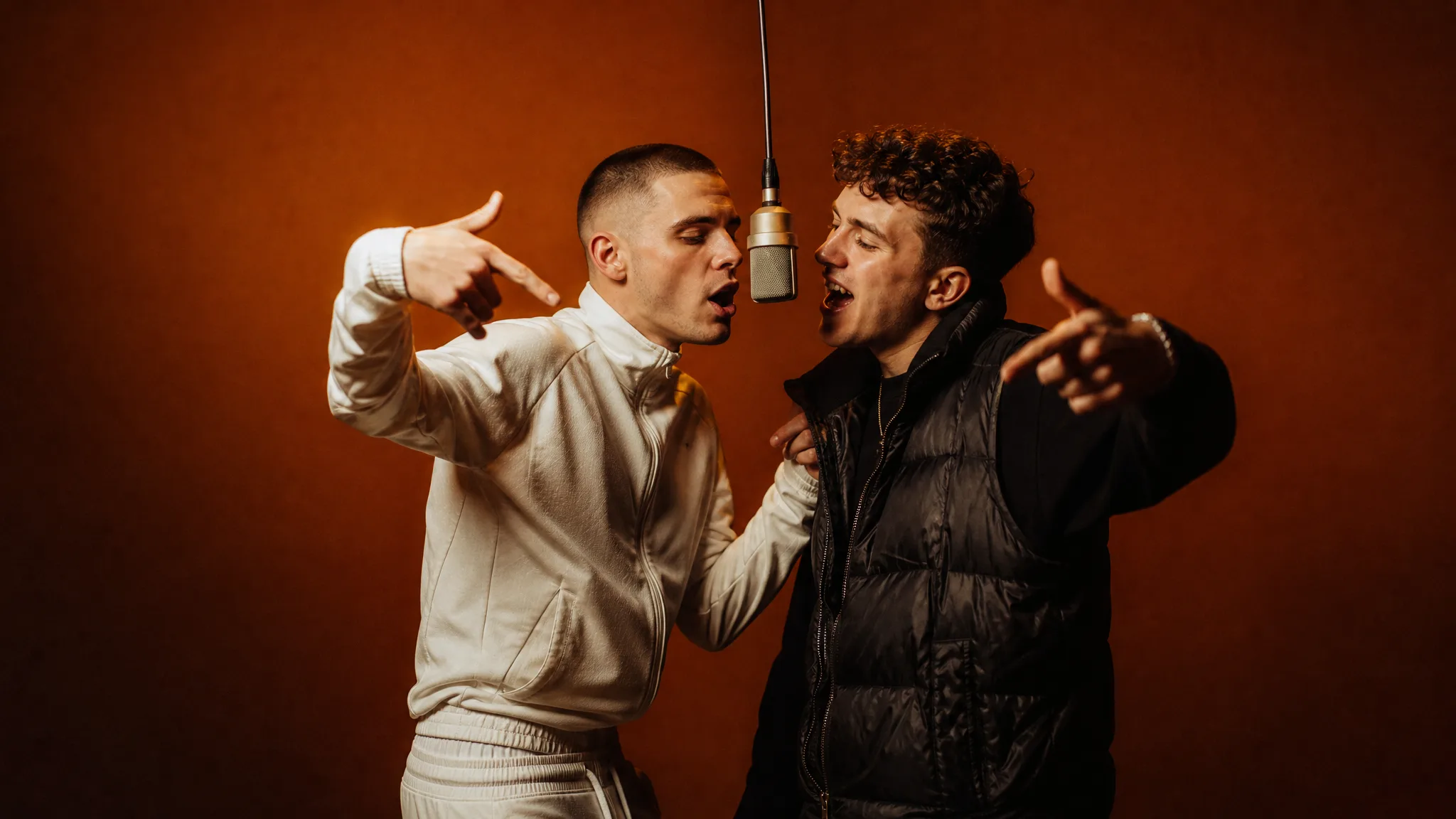 Buzz-cut brother in a white tracksuit and curly-haired brother in a black puffer vest leaning into one silver mic