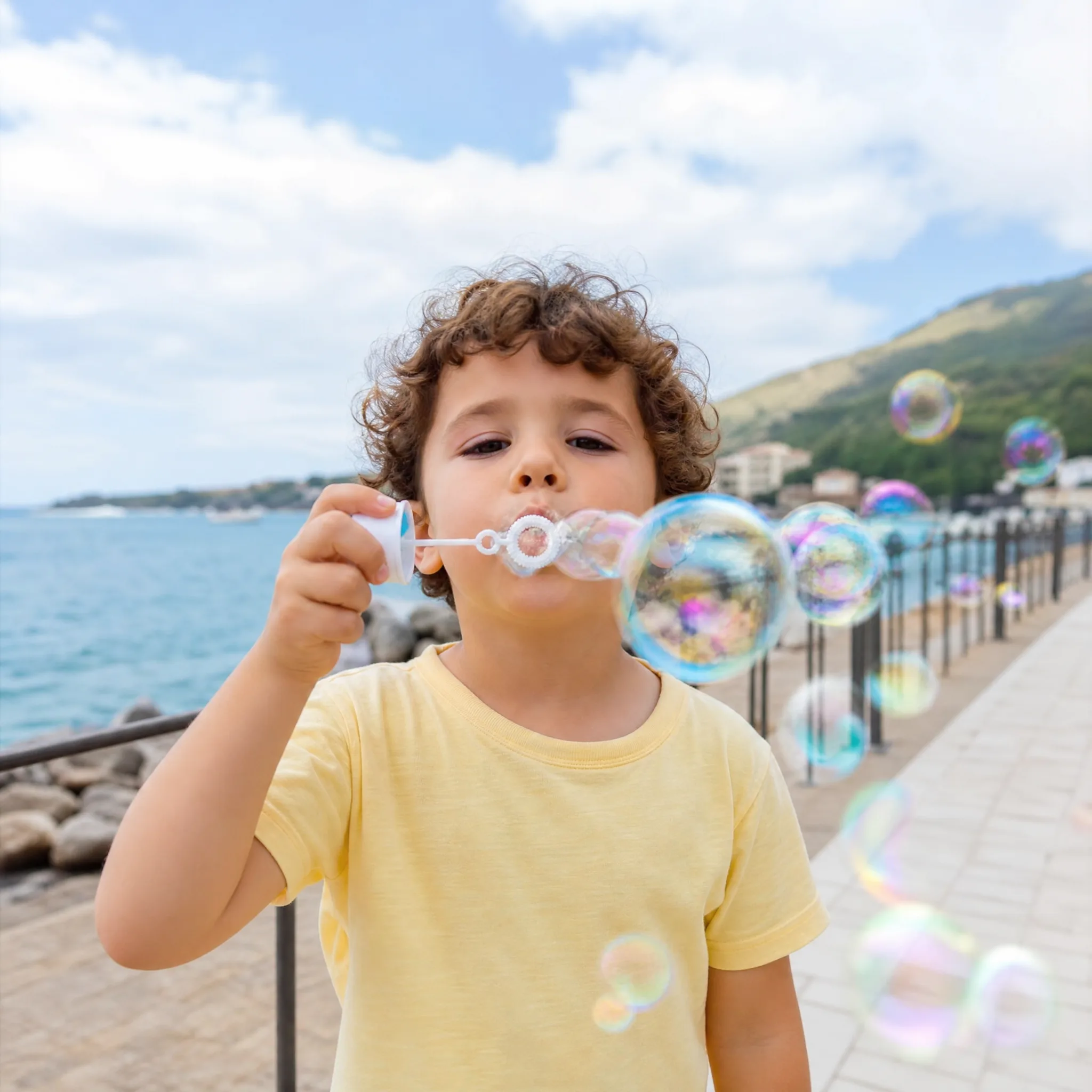 Curly-haired boy in a yellow T-shirt blows soap bubbles toward the camera on a breezy seaside promenade