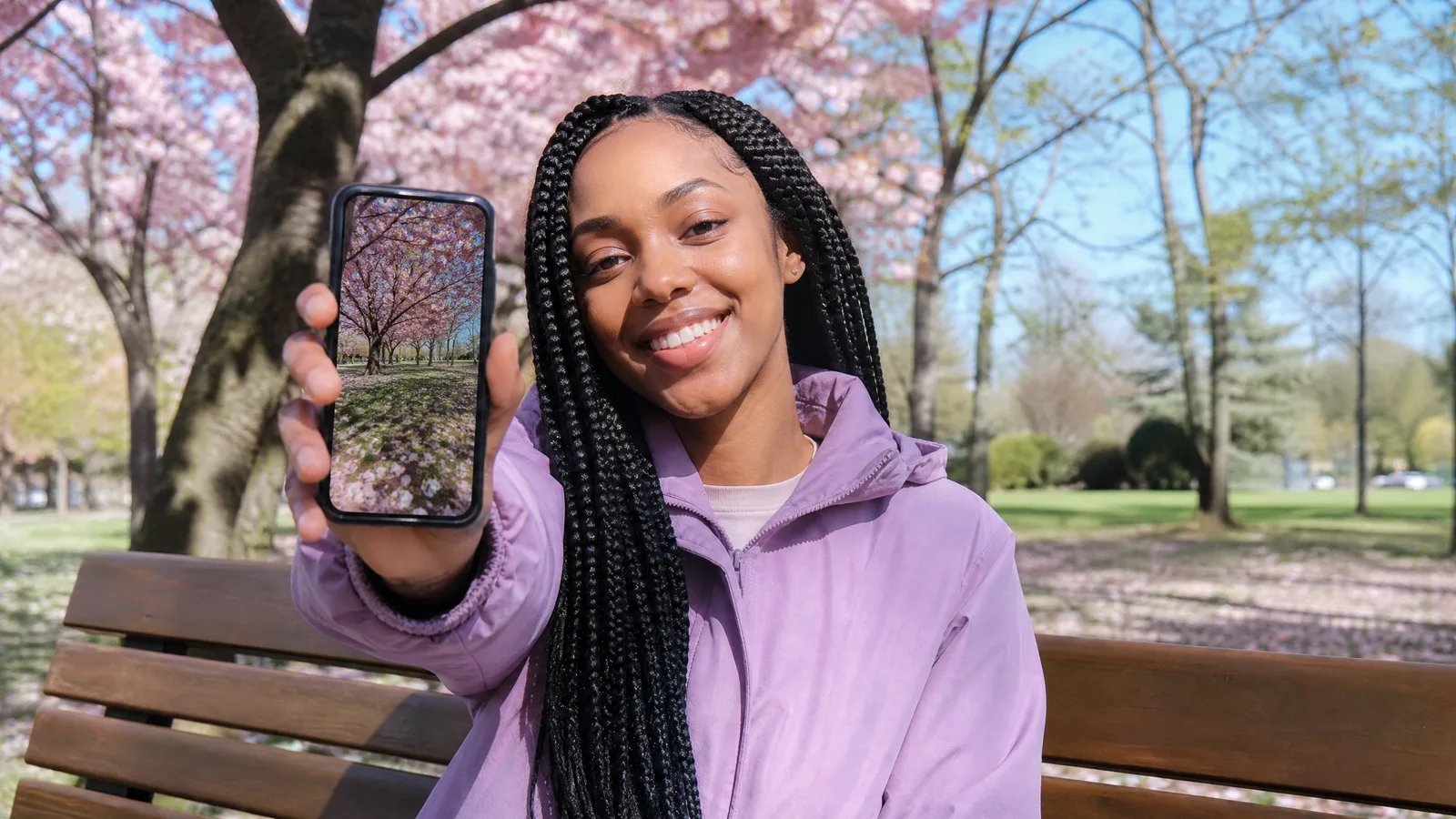A woman with box braids on a park bench holds up her phone showing the cherry blossom photo she just took