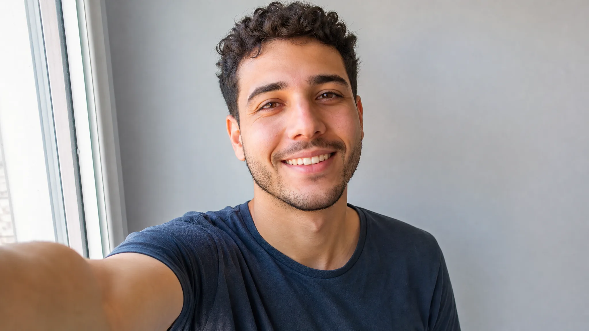 Bulin 47 AI Video Generator: Starting from a Selfie Curly-haired young man smiling into his phone for a front-facing selfie by a window, grey wall behind