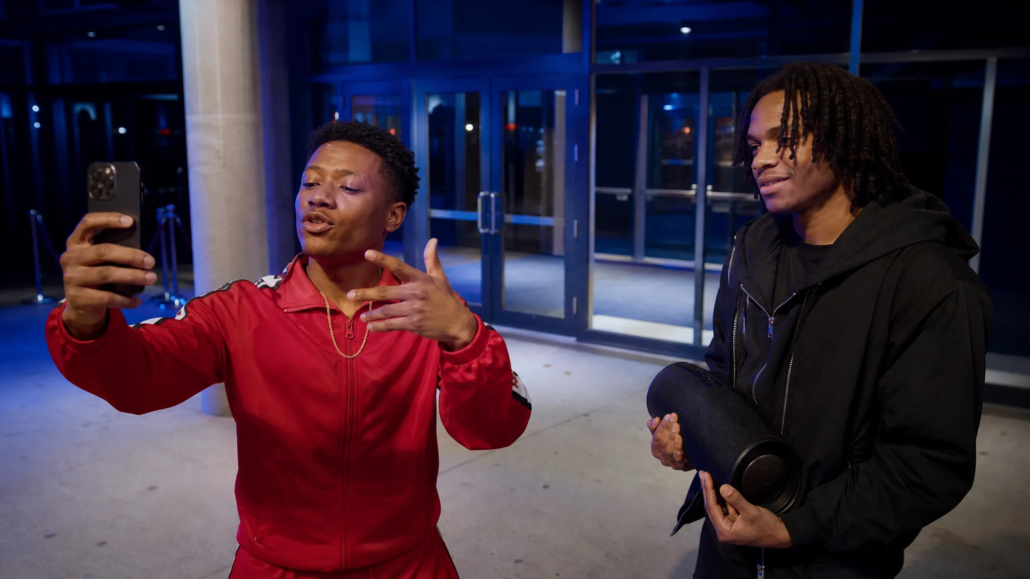 Man in a red tracksuit and gold chain rapping toward a phone at a glass venue door lit cool blue, speaker beside him
