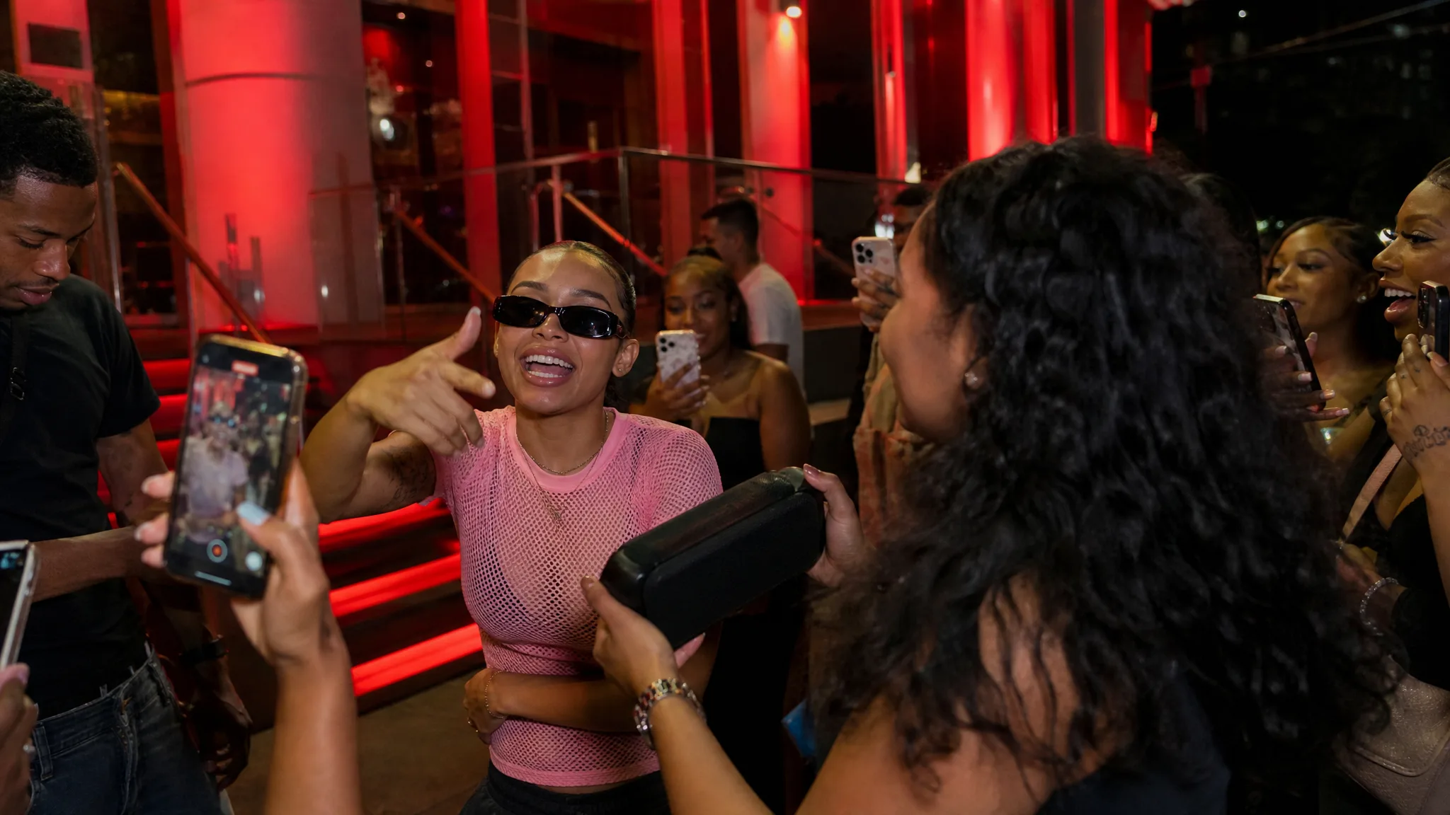 Woman in black sunglasses and a pale pink mesh shirt rapping into a phone at a red-lit entrance, a friend raising a speaker