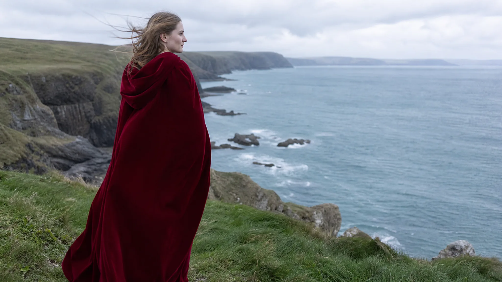 Woman in a long crimson hooded cape on a grassy sea cliff under an overcast sky, looking out over a grey-blue bay