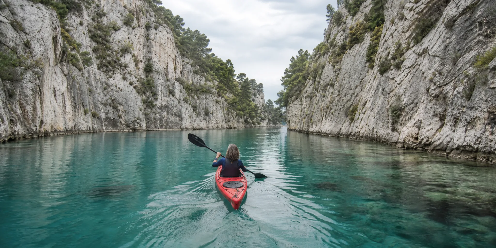 Red kayak gliding through a narrow turquoise canyon under pale limestone walls and pine trees, viewed from behind