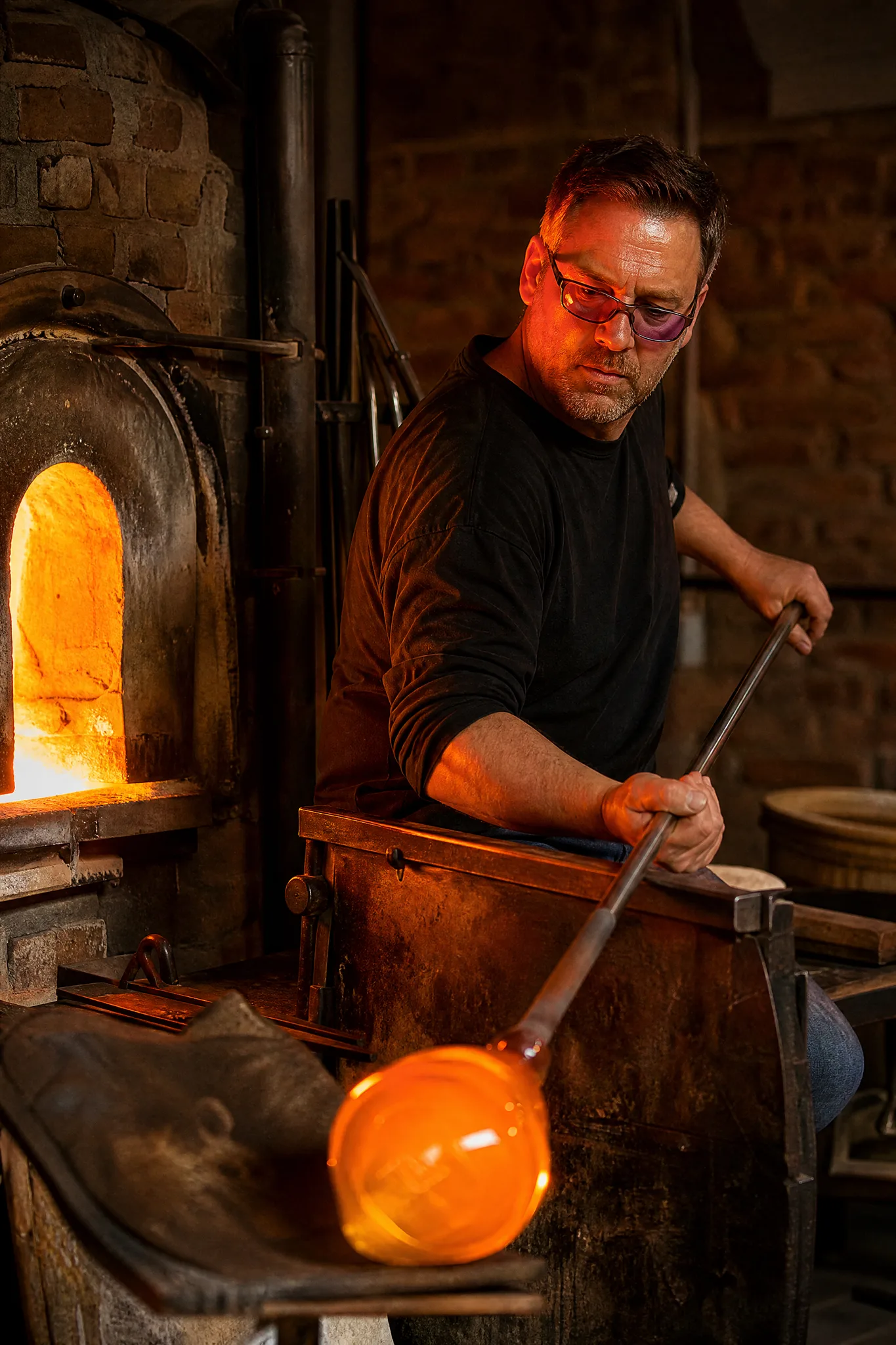 Grok Imagine Image 2: Glass at the Furnace Glassblower rotating a glowing gather of molten glass on a steel pipe next to the furnace