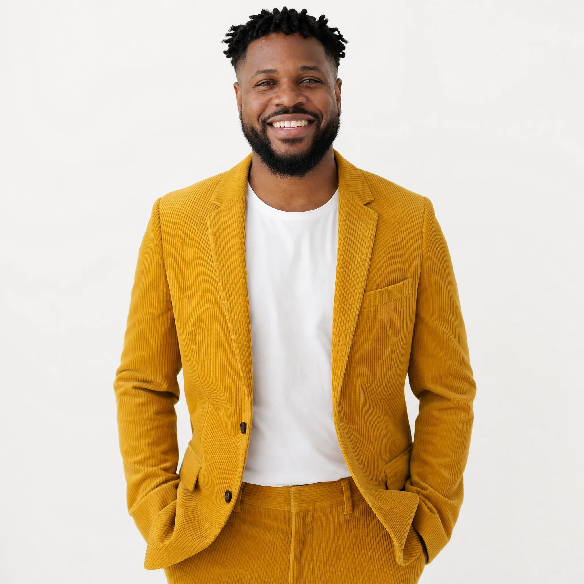 Smiling man in a mustard corduroy suit and white tee, hands in pockets, against a white studio backdrop