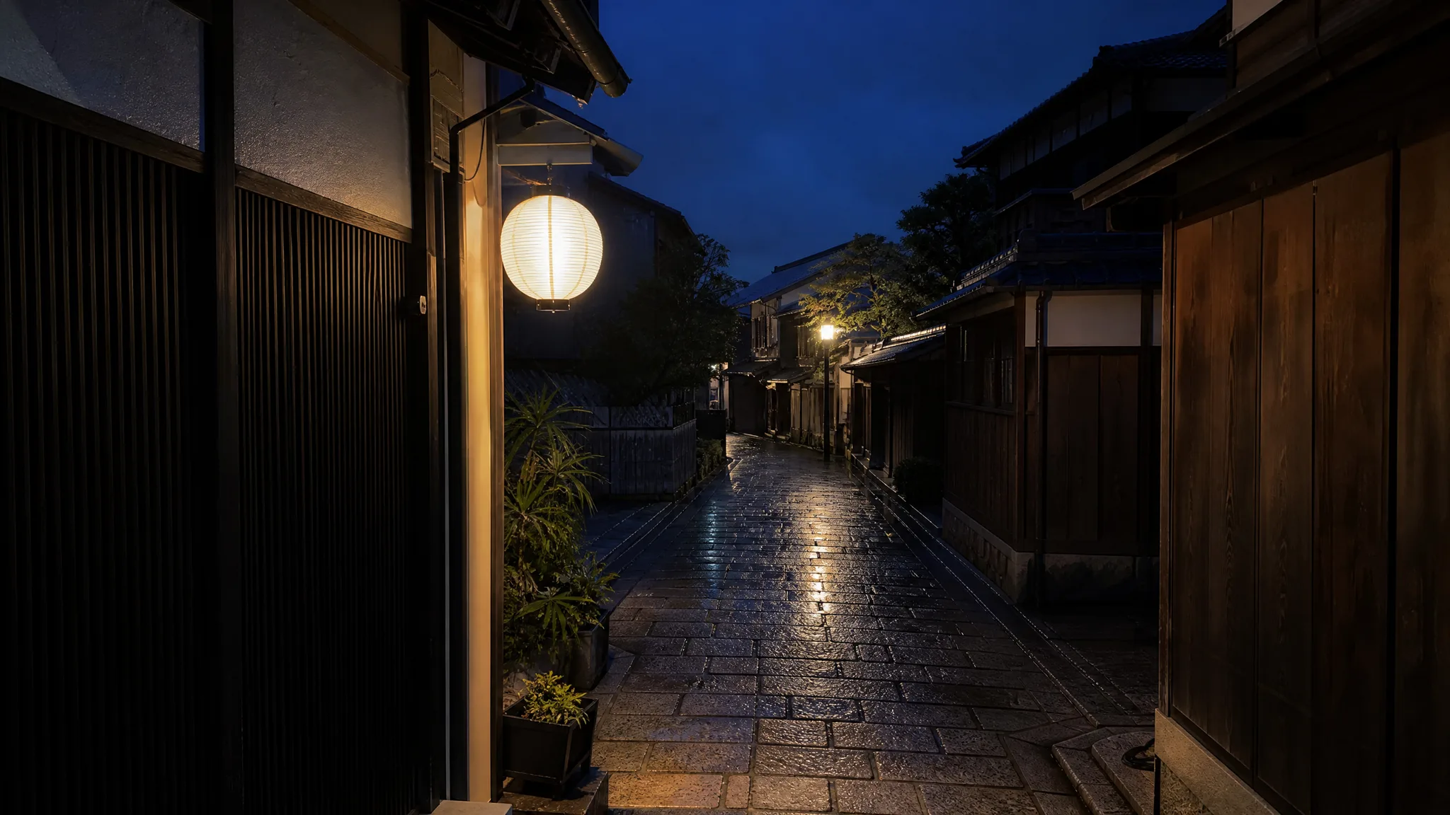 A white paper lantern glows over a wet stone alley between dark wooden houses in Kyoto under a blue night sky