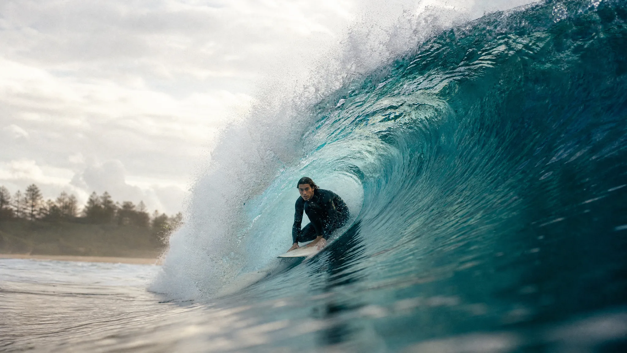 Surfer in a black wetsuit crouches inside a curling teal wave, shot from the water on a bright overcast morning