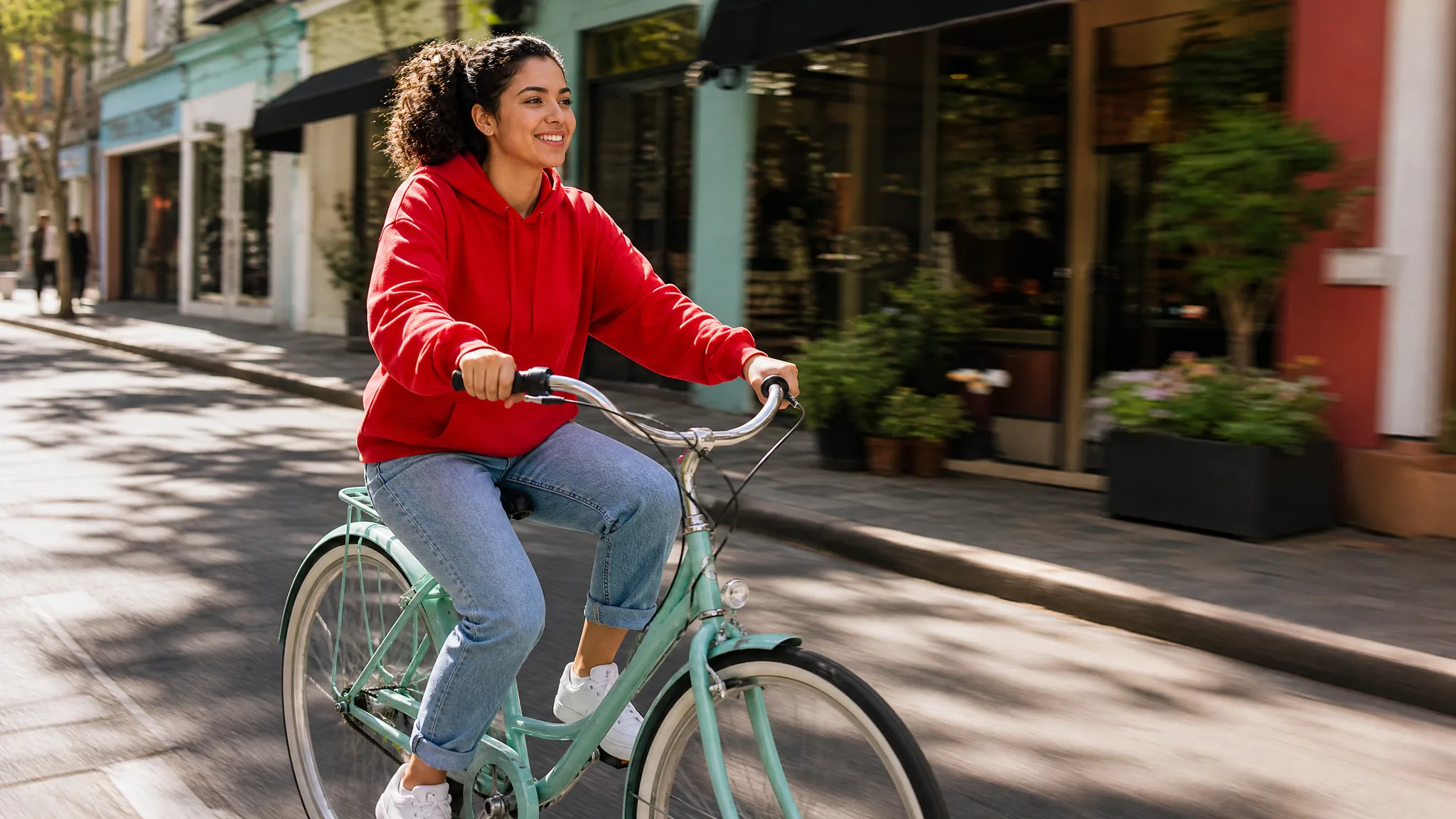 Smiling woman in a red hoodie rides a mint-green bicycle past colourful shopfronts on a sunny street