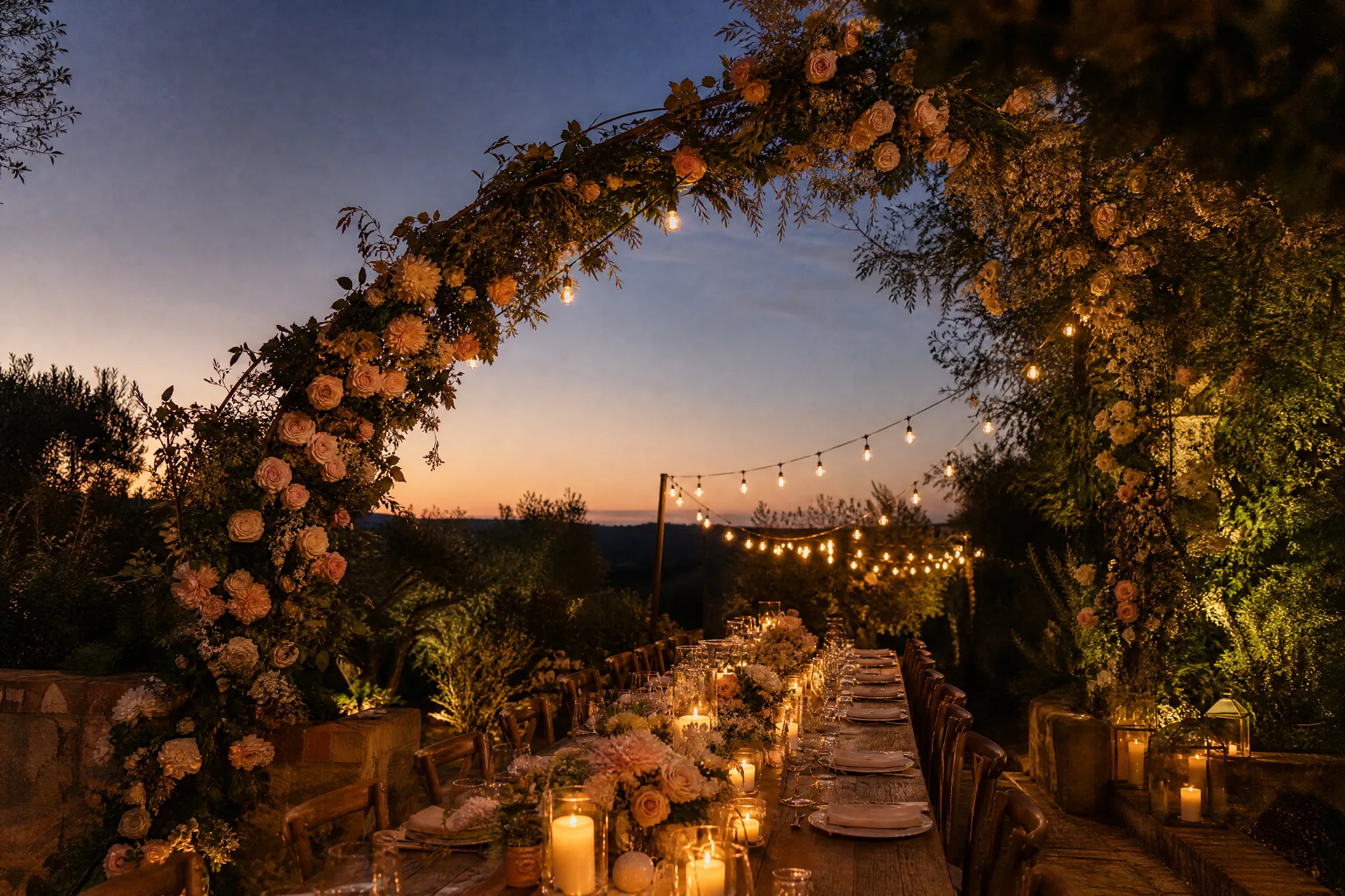 Long candlelit garden table under an arch of blush roses at dusk, with open sky above