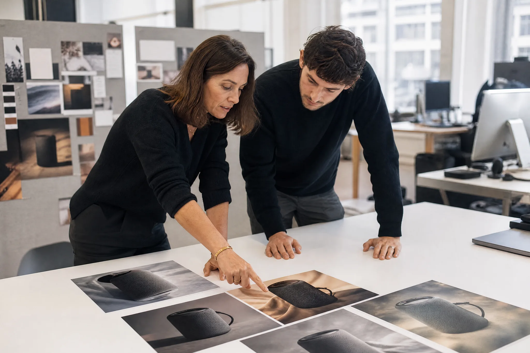 Choosing a Campaign Direction Two colleagues at a bright office table comparing printed photos of a charcoal portable speaker in different moods