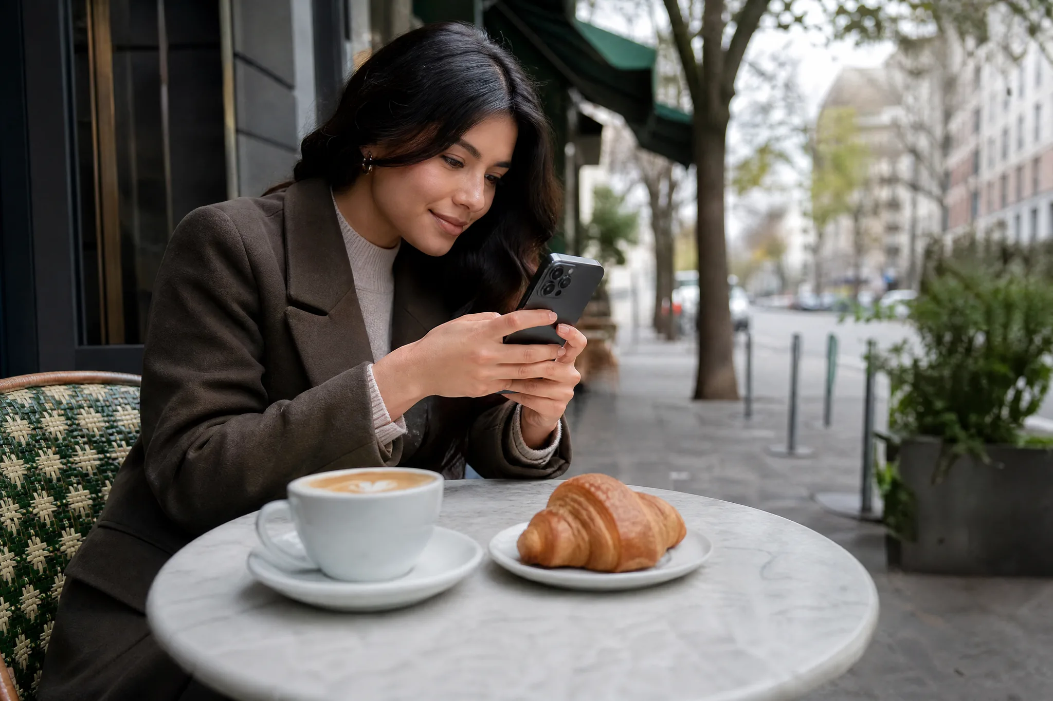 Image to Reel: A Café Photo for Your Next Post Creator at an outdoor café table on a grey morning, phone held vertically to photograph a latte and croissant