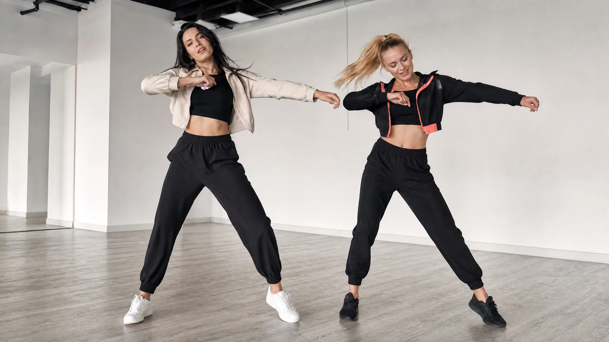 Two women dancing in a bright studio, one in a cream jacket and one in black with coral trim