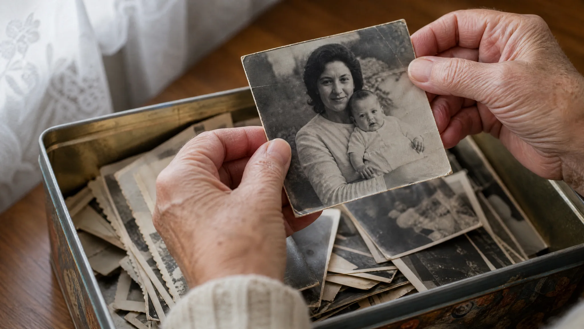 Older hands lift a black-and-white 1960s photo of a young mother and her baby from a tin full of family prints