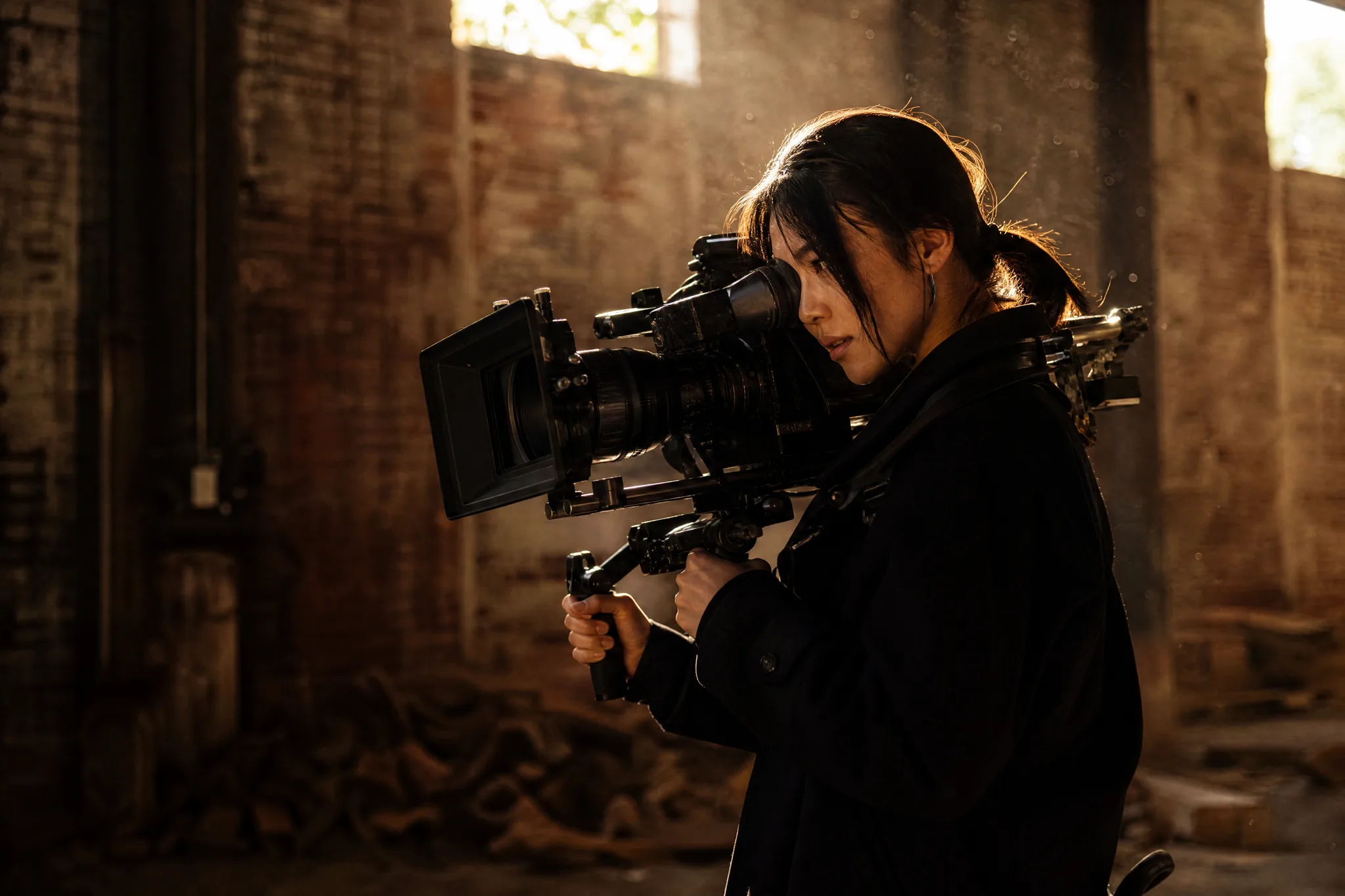 Cinematographer leaning into the viewfinder of a shoulder-rig camera as sunlight and floating dust fill a brick warehouse