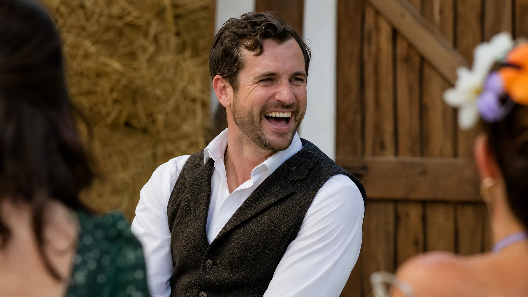 A smiling man in a brown vest watching a performance beside straw bales