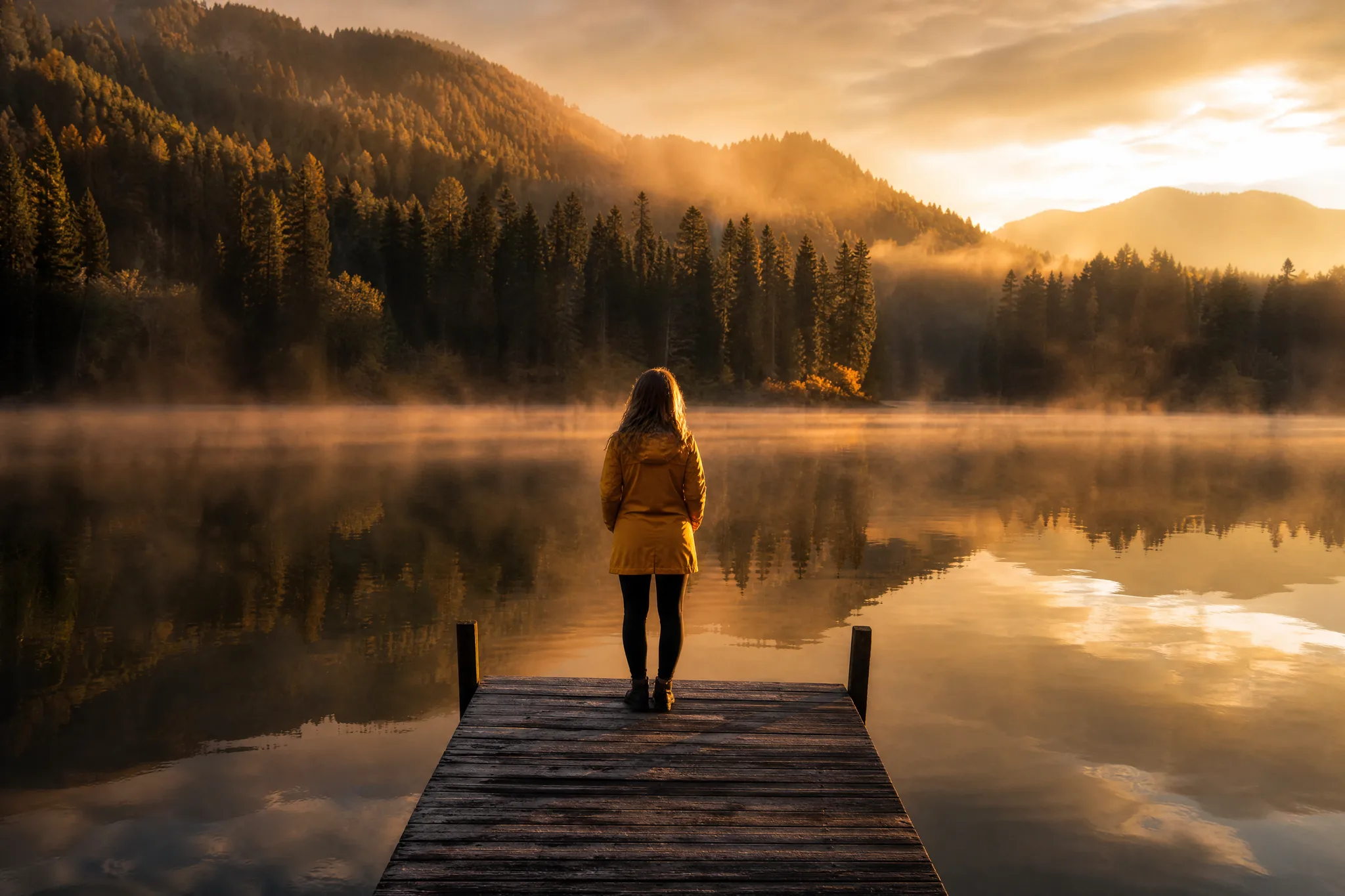 Woman in a yellow raincoat at the end of a wooden jetty on a misty lake as dawn breaks