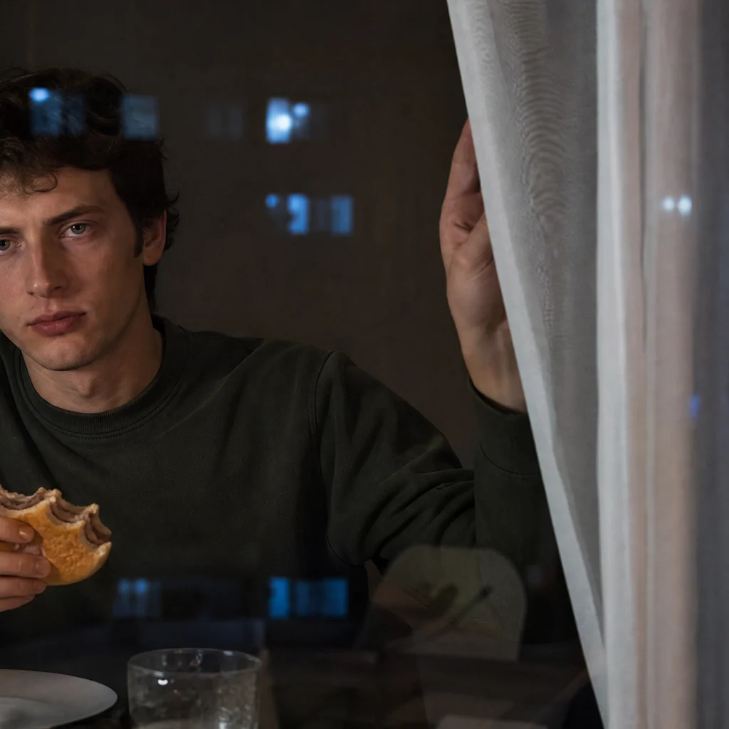 A young man holding a burger and looking out through an apartment window